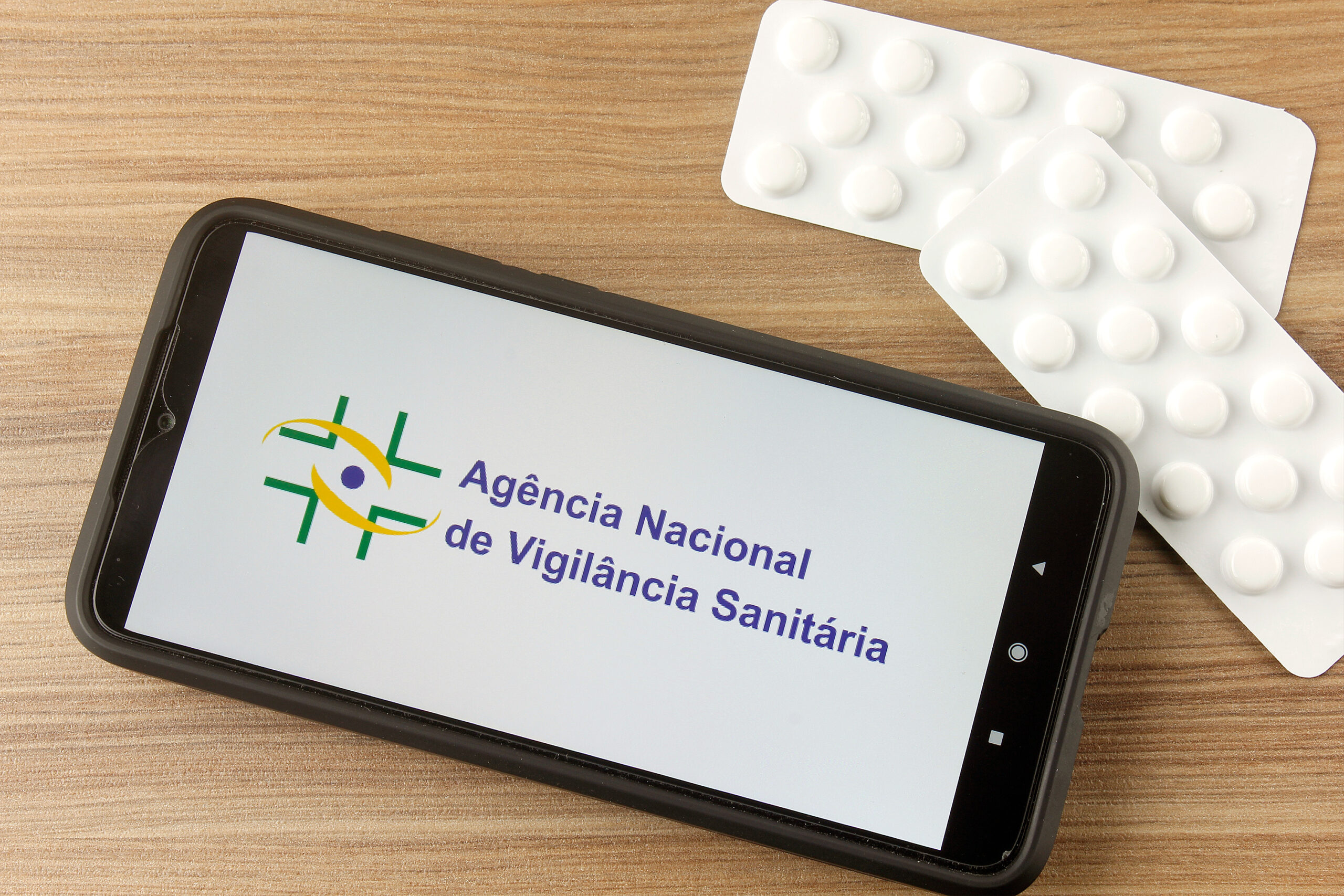 Dispositivo móvel em superfície plana ao lado de cartelas de comprimidos destaca o nome Agência Nacional de Vigilância Sanitária (ANVISA)